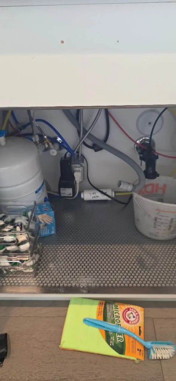 Under-sink area showing an installed reverse osmosis water filtration system with storage tank, tubing, and connections, alongside household cleaning supplies.