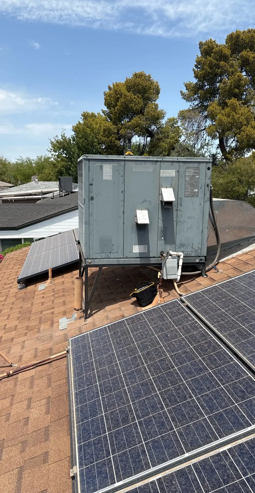 Rooftop packaged HVAC unit installed near solar panels on a shingled roof under a blue sky.