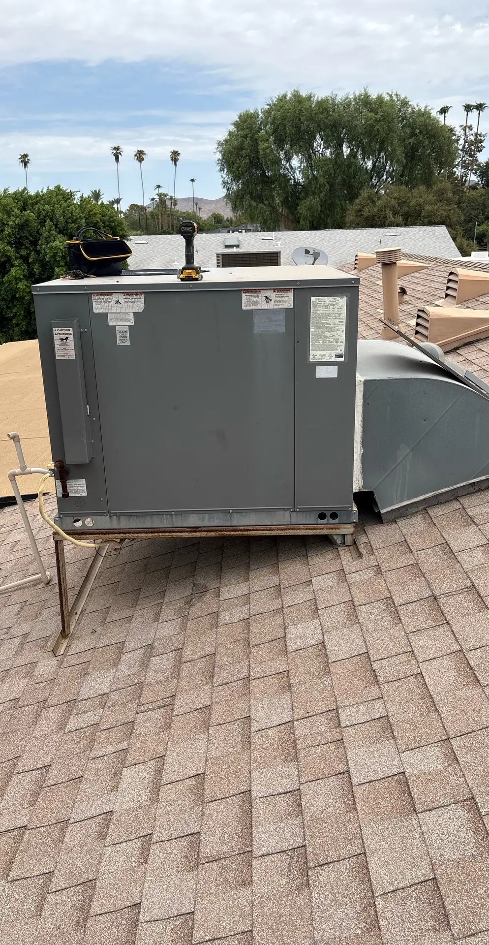 A rooftop packaged HVAC unit on shingles with tools on top and a distant residential neighborhood in the background.