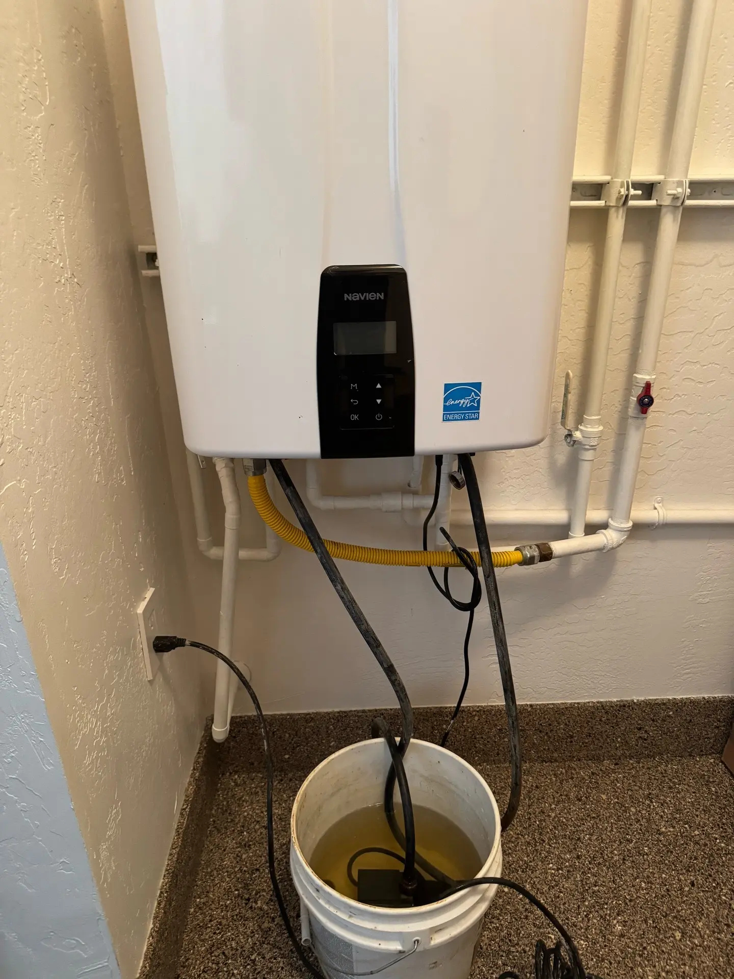 A Navien tankless water heater with descaling hoses connected, circulating cleaning solution into a bucket during a maintenance flush.