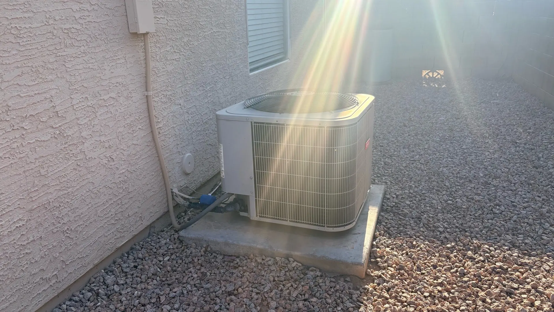 Outdoor air conditioning condenser unit on a concrete pad beside a stucco wall with gravel landscaping and sun flare.