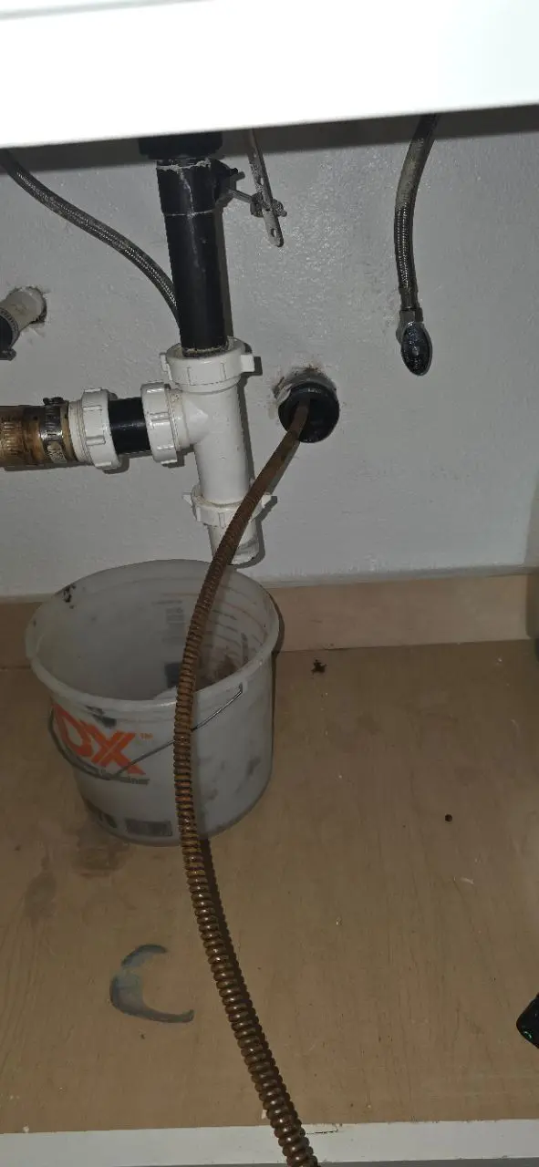 Under-sink plumbing with a drain auger cable fed into the pipe and a bucket positioned below to catch water during a drain clearing.