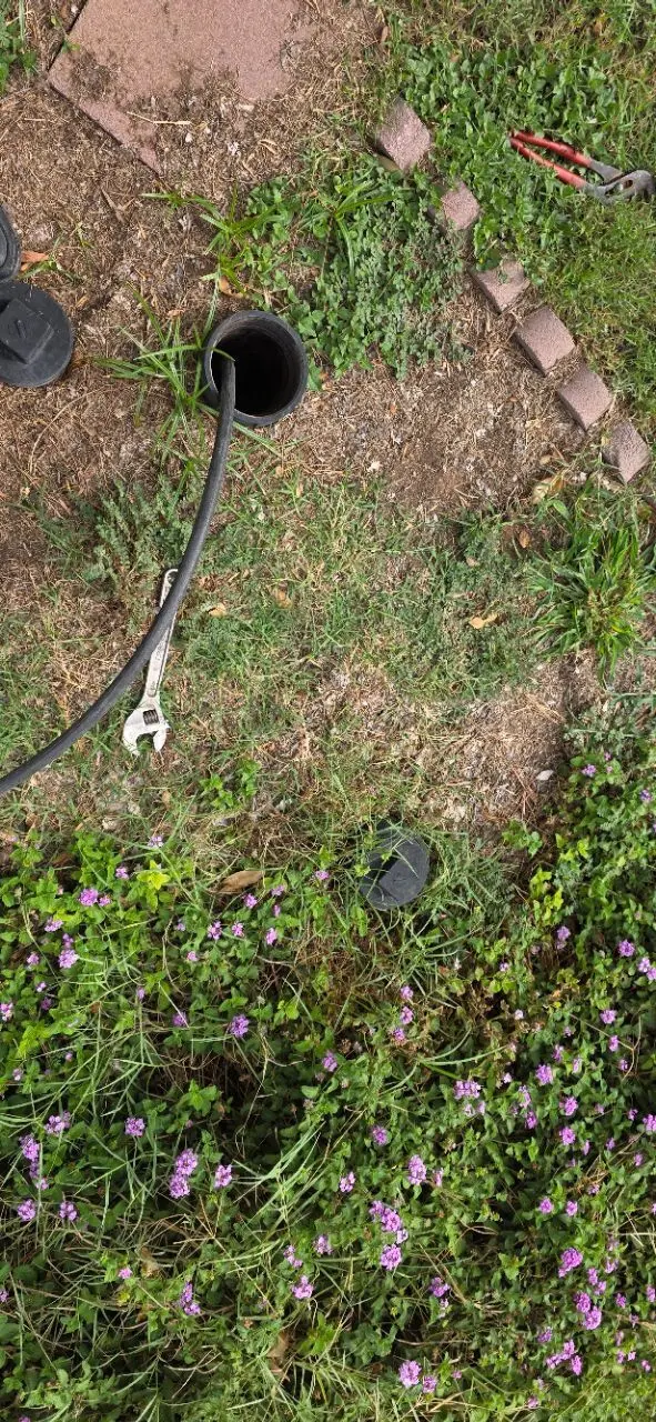 Open cleanout pipe in a grassy area with a hose and pipe wrench nearby surrounded by flowers.