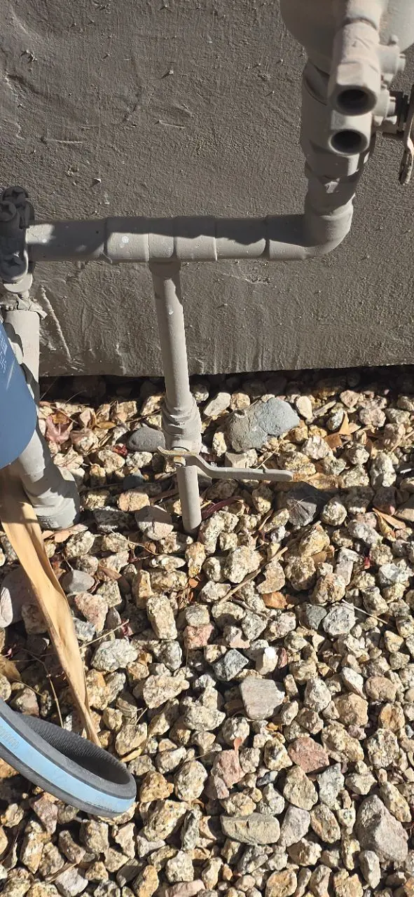 Exterior gray-painted plumbing pipes and valves against a stucco wall above landscaping gravel.},{